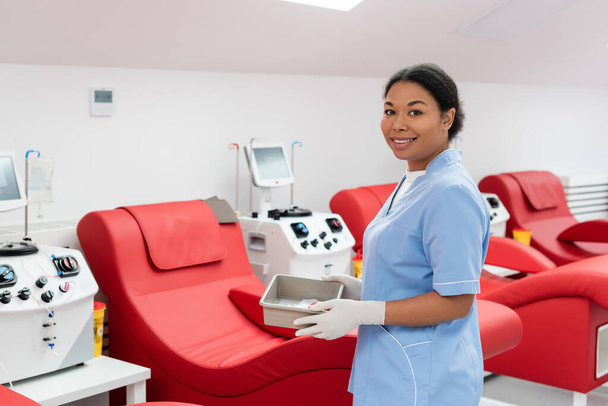 operatore sanitario multirazziale in uniforme blu e guanti in lattice con vassoio medico vicino a macchine trasfusionali e comode sedie mediche nel centro di donazione di sangue - Foto, immagini