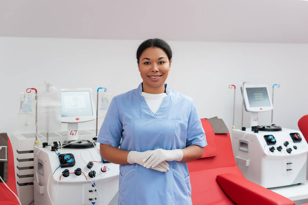 alegre enfermera multirracial en uniforme azul y guantes de látex mirando a la cámara cerca de máquinas de transfusión automatizadas y sillas médicas ergonómicas en el centro de donación de sangre - Foto, Imagen