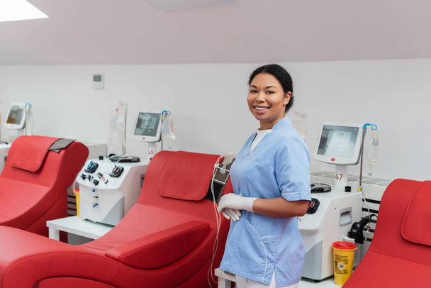 alegre enfermera multirracial en uniforme azul y guantes de látex mirando a la cámara cerca de sillas médicas y máquinas de transfusión con monitores en el centro de donación de sangre - Foto, Imagen
