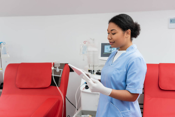 Trabajador sanitario multirracial positivo en uniforme azul y guantes de látex usando tableta digital cerca de sillas médicas y máquina de transfusión en el centro de donación de sangre - Foto, Imagen