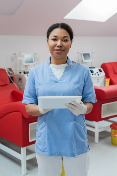 zadowolona pielęgniarka wielorasowa w niebieskich mundurach i lateksowych rękawiczkach trzymająca cyfrową tabletkę i patrząca na kamerę w pobliżu krzeseł medycznych i maszyn transfuzyjnych na rozmytym tle w centrum krwiodawstwa - Zdjęcie, obraz
