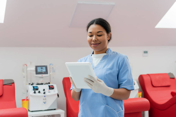 mujer multirracial sonriente en uniforme azul y guantes de látex utilizando tableta digital cerca de la máquina de transfusión automática y sillas médicas en la estación de transfusión de sangre - Foto, Imagen