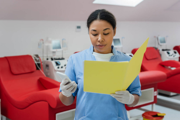 wielorasowy pracownik służby zdrowia w niebieskich mundurach i rękawicach lateksowych trzymający smartfona i czytający karty medyczne w pobliżu krzeseł medycznych i maszyn transfuzyjnych w centrum krwiodawstwa - Zdjęcie, obraz