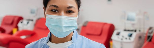 nuori monirotuinen terveydenhuollon työntekijä sinisessä univormussaan ja lääketieteellisessä naamiossa katselemassa kameraa lähellä hämärtyneitä lääketieteellisiä tuoleja ja verensiirtokoneita verenluovutuskeskuksessa, banneri - Valokuva, kuva