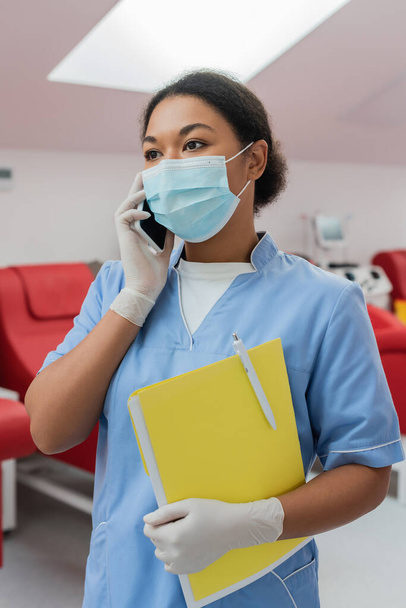 multiracial nurse in blue uniform, medical mask and latex gloves holding paper folder and pen while talking on smartphone near blurred medical chairs in blood transfusion station - Foto, Imagen
