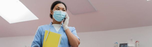 προβολή χαμηλής γωνίας της πολυφυλετικής νοσοκόμας με μπλε στολή, ιατρική μάσκα και γάντι λατέξ κρατώντας χάρτινο φάκελο με στυλό και μιλώντας στο κινητό τηλέφωνο στο κέντρο αιμοδοσίας, πανό - Φωτογραφία, εικόνα