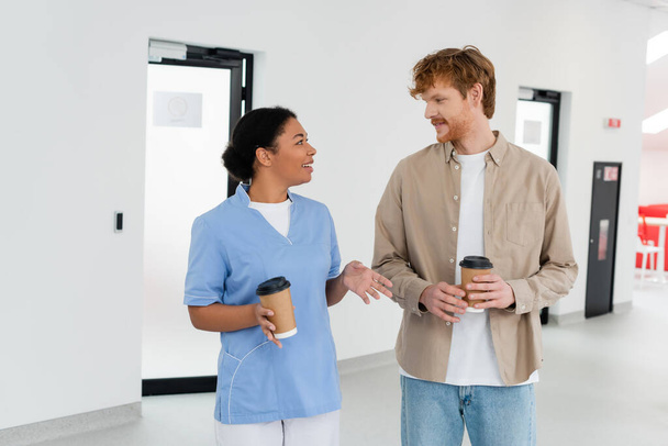 νεαρή κοκκινομάλλα εθελοντής και χαμογελαστός πολυφυλετικός εργαζόμενος υγειονομικής περίθαλψης με στολή κρατώντας κύπελλα χαρτί, ενώ μιλάμε στην αίθουσα αναμονής του κέντρου αιμοδοσίας  - Φωτογραφία, εικόνα