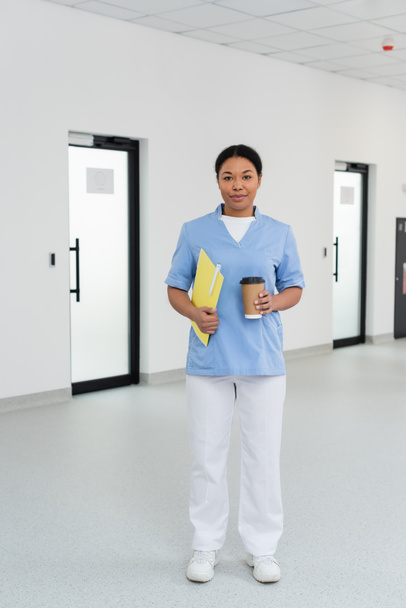 full length of young and smiling multiracial nurse with paper folder, pen and takeaway coffee looking at camera in waiting area of blood donation center, door on background  - Photo, Image