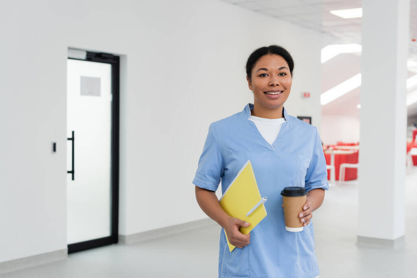 ευτυχισμένη πολυφυλετική νοσοκόμα σε μπλε στολή κρατώντας φάκελο με στυλό, κύπελλο μιας χρήσης με καφέ και χαμογελώντας στην κάμερα στην αίθουσα αναμονής του κέντρου αιμοδοσίας, πόρτα στο παρασκήνιο  - Φωτογραφία, εικόνα