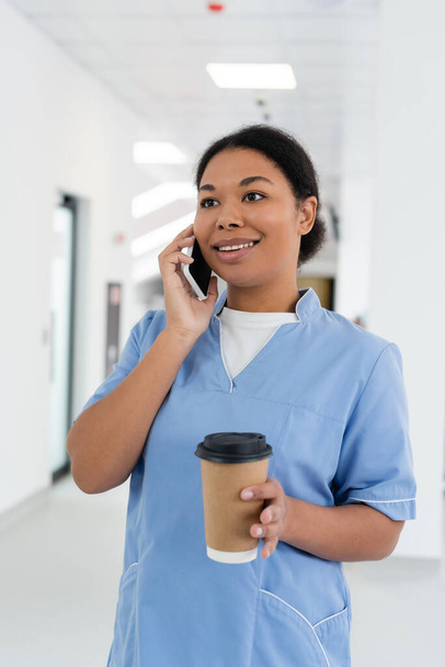 enfermera multirracial positiva sonriendo durante la conversación en el teléfono inteligente, mientras que de pie con café para llevar en el pasillo de la estación de transfusión de sangre, sala borrosa en el fondo  - Foto, imagen