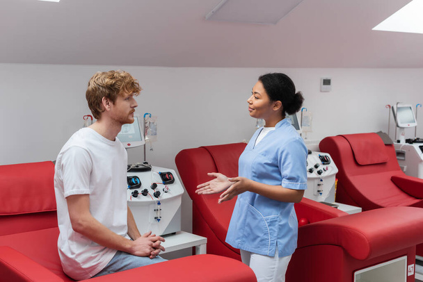 enfermeira multirracial em uniforme azul gestos e conversando com doador de sangue jovem ruiva sentado na cadeira médica perto de máquinas de transfusão no hospital - Foto, Imagem