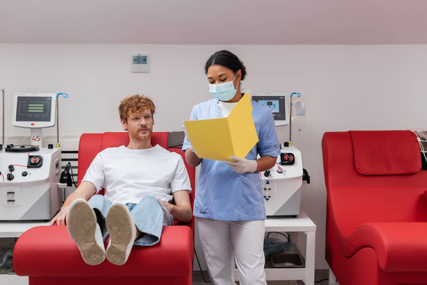 multiracial healthcare worker in uniform and medical mask showing paper folder to redhead volunteer sitting on medical chair near transfusion machine in laboratory - Photo, Image