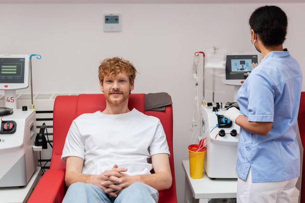 πολυφυλετική νοσοκόμα σε μπλε στολή λειτουργίας της μηχανής μετάγγισης κοντά σε πλαστικό κύπελλο και κοκκινομάλλα εθελοντής κάθεται στην ιατρική καρέκλα και κοιτάζοντας κάμερα στο κέντρο αιμοδοσίας - Φωτογραφία, εικόνα