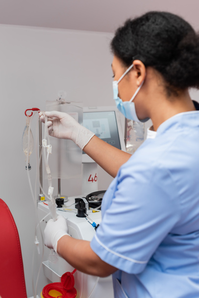 multiraciale verpleegkundige in blauw uniform en latex handschoenen met bloedtransfusieset in de buurt van infuusstandaard met infuuszakken in laboratorium, wazig voorgrond - Foto, afbeelding
