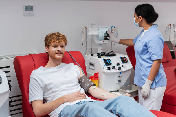 multiracial nurse in latex gloves and medical mask operating transfusion machine near positive redhead volunteer sitting on comfortable chair in blood donation center - Фото, изображение