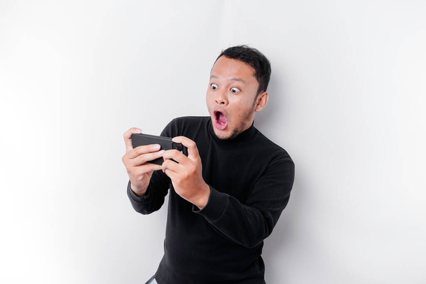 Young Asian man in casual wear is playing games with mobile phone with a white background isolated. - Photo, image