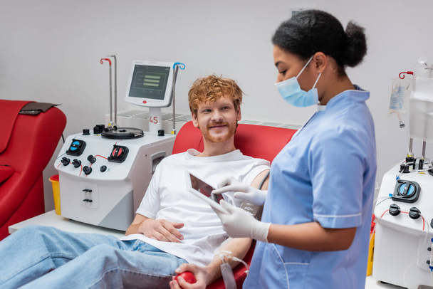 πολυφυλετική νοσοκόμα με μπλε στολή και ιατρική μάσκα στέκεται με ψηφιακή ταμπλέτα κοντά χαμογελαστή κοκκινομάλλα εθελοντής σε άνετη καρέκλα δίπλα σε αυτοματοποιημένο εξοπλισμό στο κέντρο αιμοδοσίας - Φωτογραφία, εικόνα