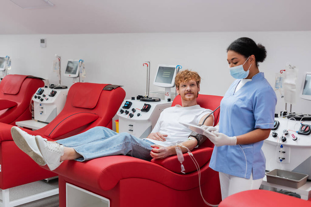 multiraciális nővér orvosi maszkban és latex kesztyű digitális tabletta közelében vörös hajú férfi vérátömlesztés készlet ül kényelmes ergonomikus szék mellett automatizált berendezések klinikán - Fotó, kép