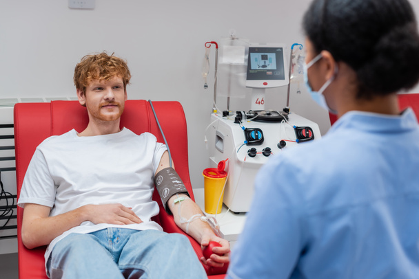 輸血機の近くの医療椅子に座っている間にゴムボールを保持している血圧カフの赤毛の男、前景にぼやけた額に医療マスクでプラスチックカップと多人種看護師 - 写真・画像
