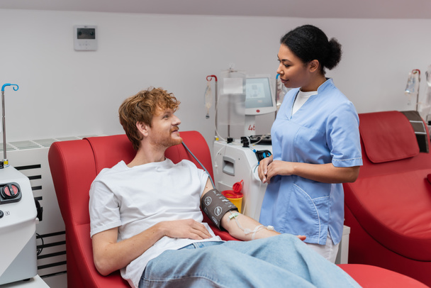 többfunkciós nővér kék egyenruhában beszél vörös hajú vérnyomásmérővel és transzfúziós készlettel, aki orvosi széken ül a véradó központ automatizált berendezéseinek közelében. - Fotó, kép