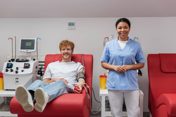 onnellinen punapää mies verenpaineen mansetti ja kumipallo istuu lääketieteellisellä tuolilla lähellä monirotuinen sairaanhoitaja, verensiirto kone ja muovi kupit laboratoriossa, katsot kameraa  - Valokuva, kuva