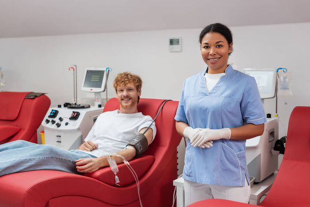 κοκκινομάλλα εθελόντρια με cuff πίεσης και σετ μετάγγισης χαμογελώντας στην κάμερα κοντά σε πολυφυλετική νοσοκόμα με γάντια λάτεξ και αυτοματοποιημένο εξοπλισμό στο κέντρο αιμοδοσίας - Φωτογραφία, εικόνα