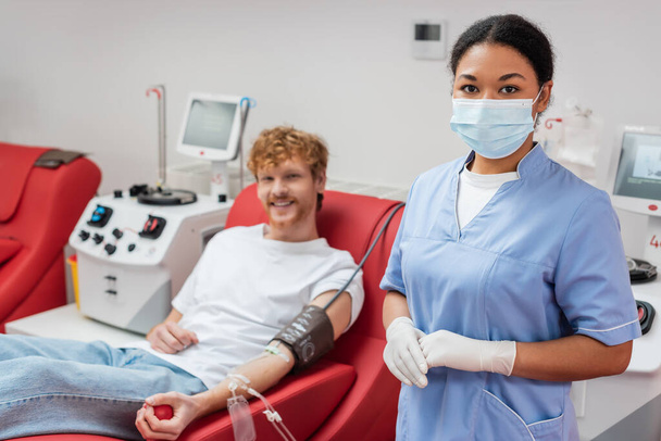 multiraciale gezondheidswerker in medisch masker en latex handschoenen die naar camera kijken bij wazige man met bloedtransfusieset zittend op ergonomische stoel naast transfusiemachines in het laboratorium - Foto, afbeelding