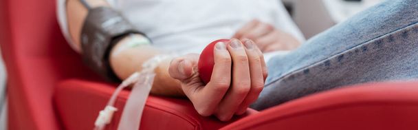 vista parcial de un joven voluntario sentado en una silla ergonómica cómoda y apretando la bola de goma durante el procedimiento médico en el centro de donación de sangre, pancarta - Foto, Imagen