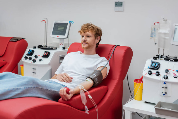 young redhead donor with blood pressure cuff and transfusion set sitting on ergonomic and comfortable medical chair near automated equipment and plastic cups in clinic  - Фото, изображение