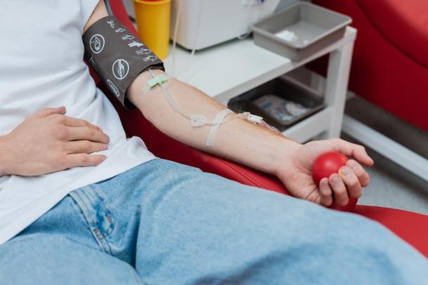 vista parcial del donante con el conjunto de transfusión apretando la bola de goma mientras está sentado en una silla cómoda cerca de la bandeja médica borrosa y la taza de plástico en el centro de donación de sangre - Foto, Imagen
