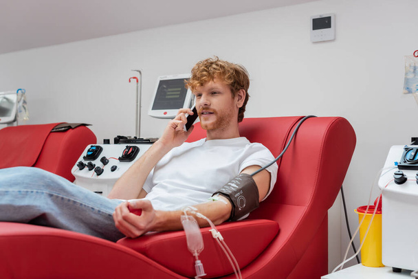 roodharige donor met transfusie set zittend op medische stoel en praten op smartphone in de buurt van moderne geautomatiseerde apparatuur en plastic beker in bloed donatie centrum - Foto, afbeelding