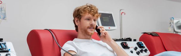 fiatal és vörös hajú önkéntes mobiltelefonnal beszél miközben vért ad orvosi székre közel a modern automatizált transzfúziós géphez a laborban, banner - Fotó, kép