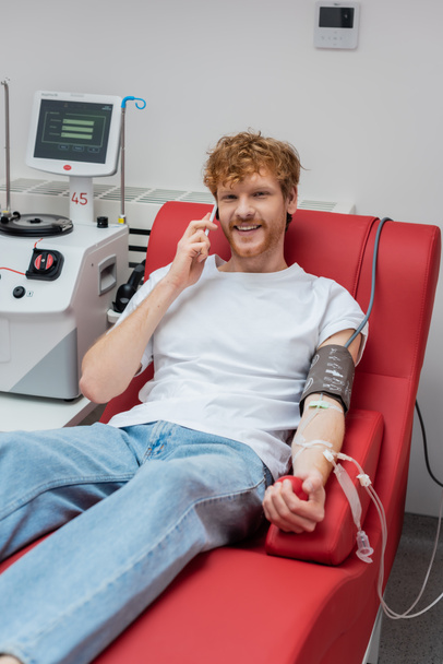 χαρούμενη κοκκινομάλλα εθελόντρια με cuff πίεσης και σετ μετάγγισης που μιλάει στο κινητό τηλέφωνο σε καρέκλα κοντά σε σύγχρονο αυτοματοποιημένο εξοπλισμό στο νοσοκομείο - Φωτογραφία, εικόνα