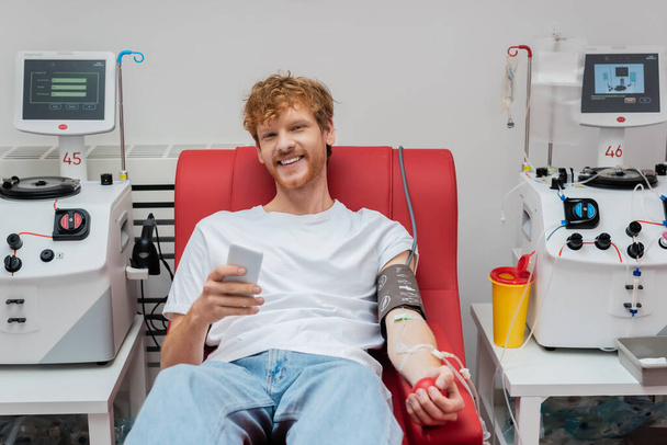 veselý zrzka muž s krevním tlakem manžety a transfuzní set drží mobilní telefon, zatímco sedí na lékařské židli v blízkosti automatizovaného zařízení a plastový kelímek v laboratoři - Fotografie, Obrázek