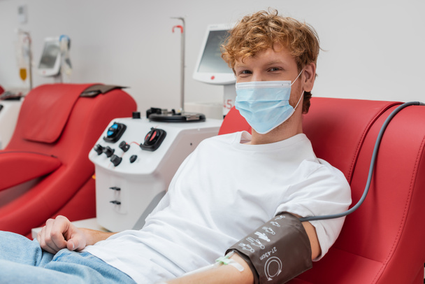 zrzka dárce v lékařské masce a krevní tlak manžety při pohledu do kamery, zatímco sedí na ergonomické židli v blízkosti automatizovaného transfuzního stroje v nemocnici - Fotografie, Obrázek