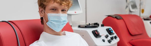 fiatal és vörös hajú önkéntes orvosi maszkban, pozitív szemkifejezéssel, vért adományoz a kortárs transzfúziós gép közelében, és kamerába néz a laborban, banner - Fotó, kép