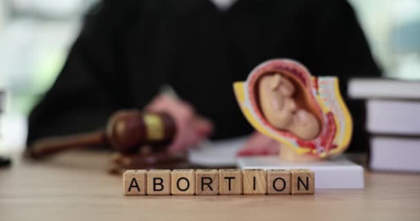 Word Abortion with wooden cubes on judge counter in office. Judge takes notes to prosecute next to fetal model. Concept legislation against abortion slow motion - Footage, Video