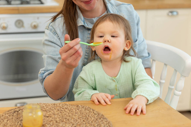 Щаслива родина вдома. Мати годує свою дитину з ложки на кухні. Маленька дитина з пухнастим смішним обличчям їсть здорову їжу вдома. Молода жінка мама дає їжу доньці
 - Фото, зображення