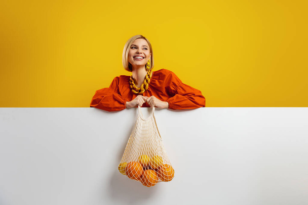 Happy young woman carrying a mesh bag with fresh oranges against yellow background - 写真・画像