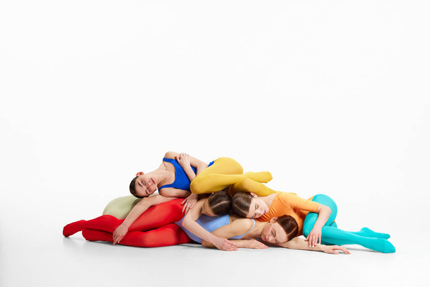 Group of tender, young girls, ballet dancers in colorful, bright clothes lying on each other on floor, dancing against grey studio background. Creativity, classic dance style, contemporary art concept - Φωτογραφία, εικόνα
