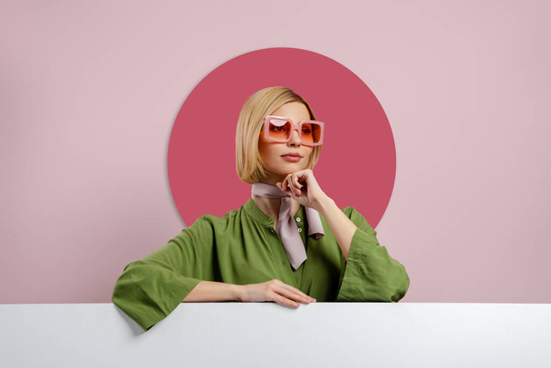 Fashionable young woman in trendy eyeglasses leaning at copy space against pink background - Zdjęcie, obraz