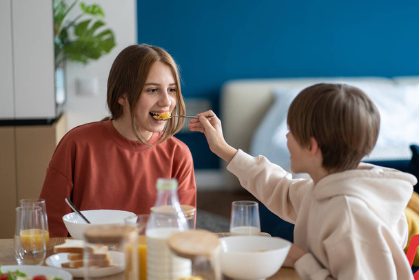 Boldog gyerekek testvérek szórakoznak együtt eszik gabonapelyhet reggel ül a konyhaasztalnál, testvér etetés testvér kukorica pelyhek. Európai családi kezdőnap egészséges reggelivel, étkezés közben - Fotó, kép