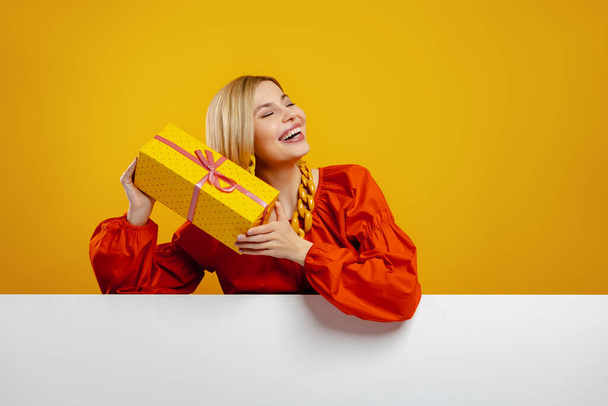 Curious young woman holding a gift box near face and smiling against yellow background - Fotografie, Obrázek