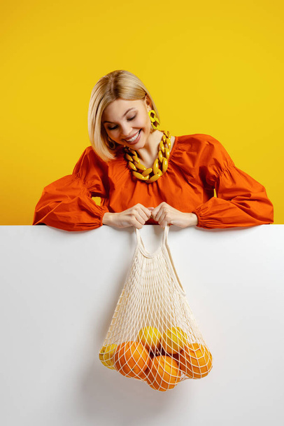 Attractive young woman looking at the mesh bag with oranges against yellow background - Fotografie, Obrázek