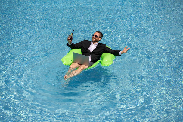 Successful business man floating in suit in pool water. Summer vacations and travel concept. Funny business man drink summer cocktail and using laptop in swimming pool. Summer business concept - Foto, imagen