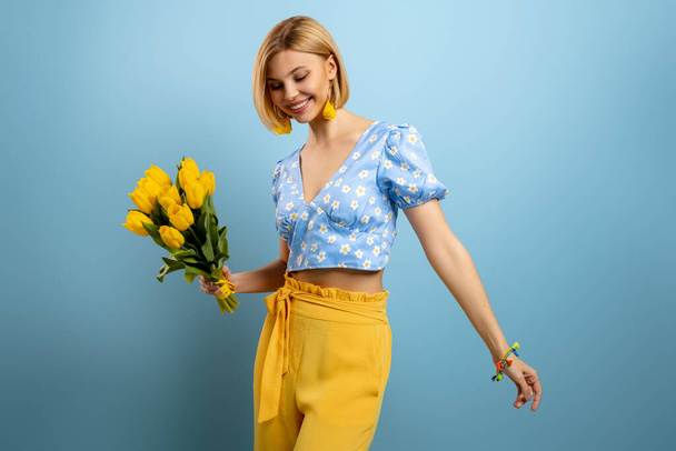 Happy young woman holding bunch of yellow tulips against blue background - Fotografie, Obrázek