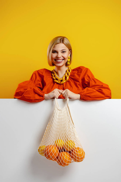 Happy young woman carrying a mesh bag with fresh oranges against yellow background - Foto, afbeelding