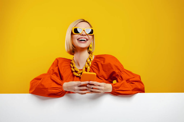 Fashionable young woman in trendy glasses holding smart phone against yellow background - Valokuva, kuva