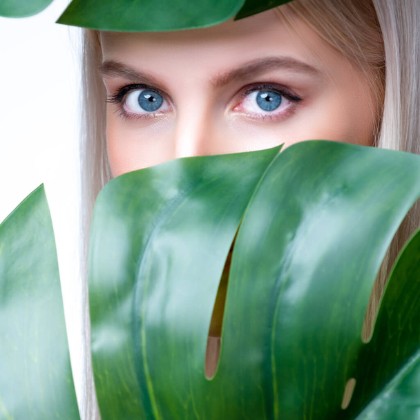 Closeup facial portrait personable woman with perfect smooth makeup holding green monstera leaves and cover her face as natural healthy skincare treatment. Tropical nature and beauty concept - Fotoğraf, Görsel