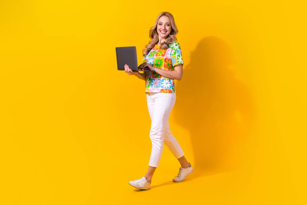Full length φωτογραφία του όμορφου κοριτσιού ντυμένο πολύχρωμο t-shirt λευκό παντελόνι κρατήσει φορητό υπολογιστή με τα πόδια κενό χώρο που απομονώνονται σε κίτρινο χρώμα φόντο. - Φωτογραφία, εικόνα